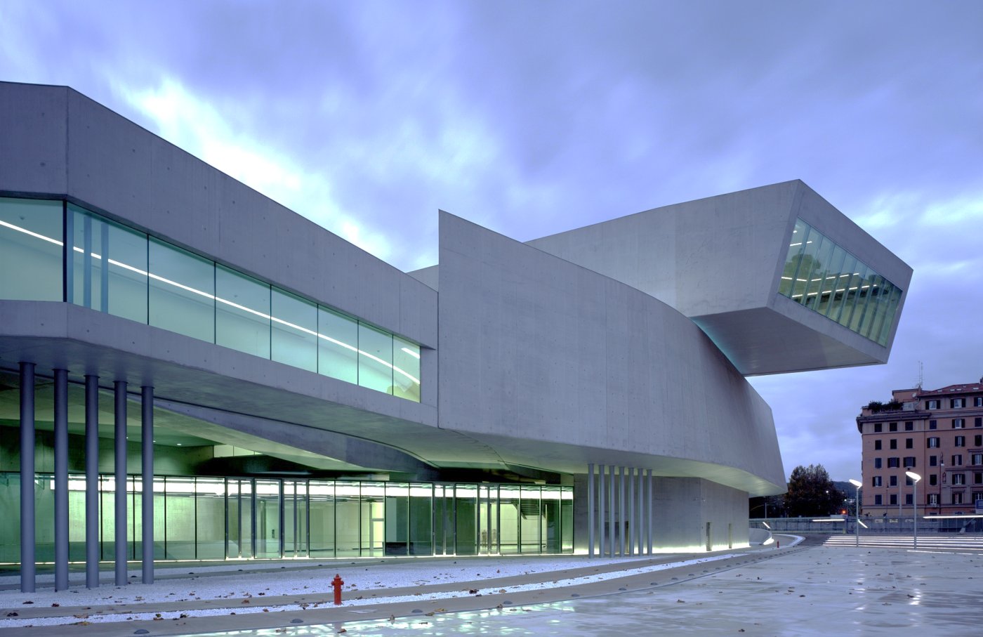 Long concrete and glass volumes cantilevering over an open forecourt at dusk