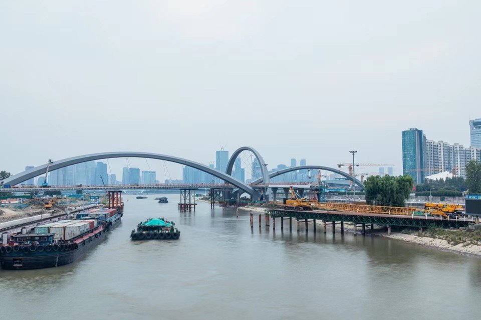 Arches of a bridge under construction spanning a wide river, with working barges passing beneath
