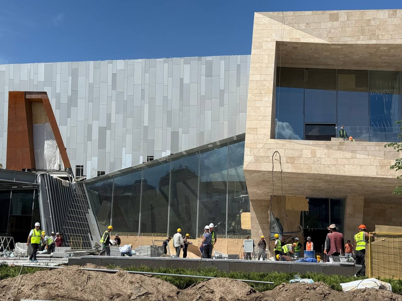 A stone-clad museum under construction with a glazed corner, workers on the ground in front of it