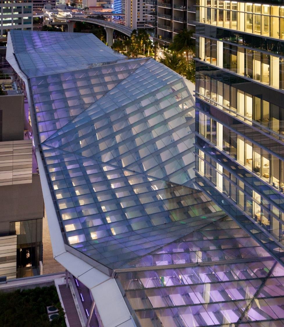 Glass above, fins across. The climate ribbon keeps out rain and sun while catching the prevailing winds and directing them into the arcade.