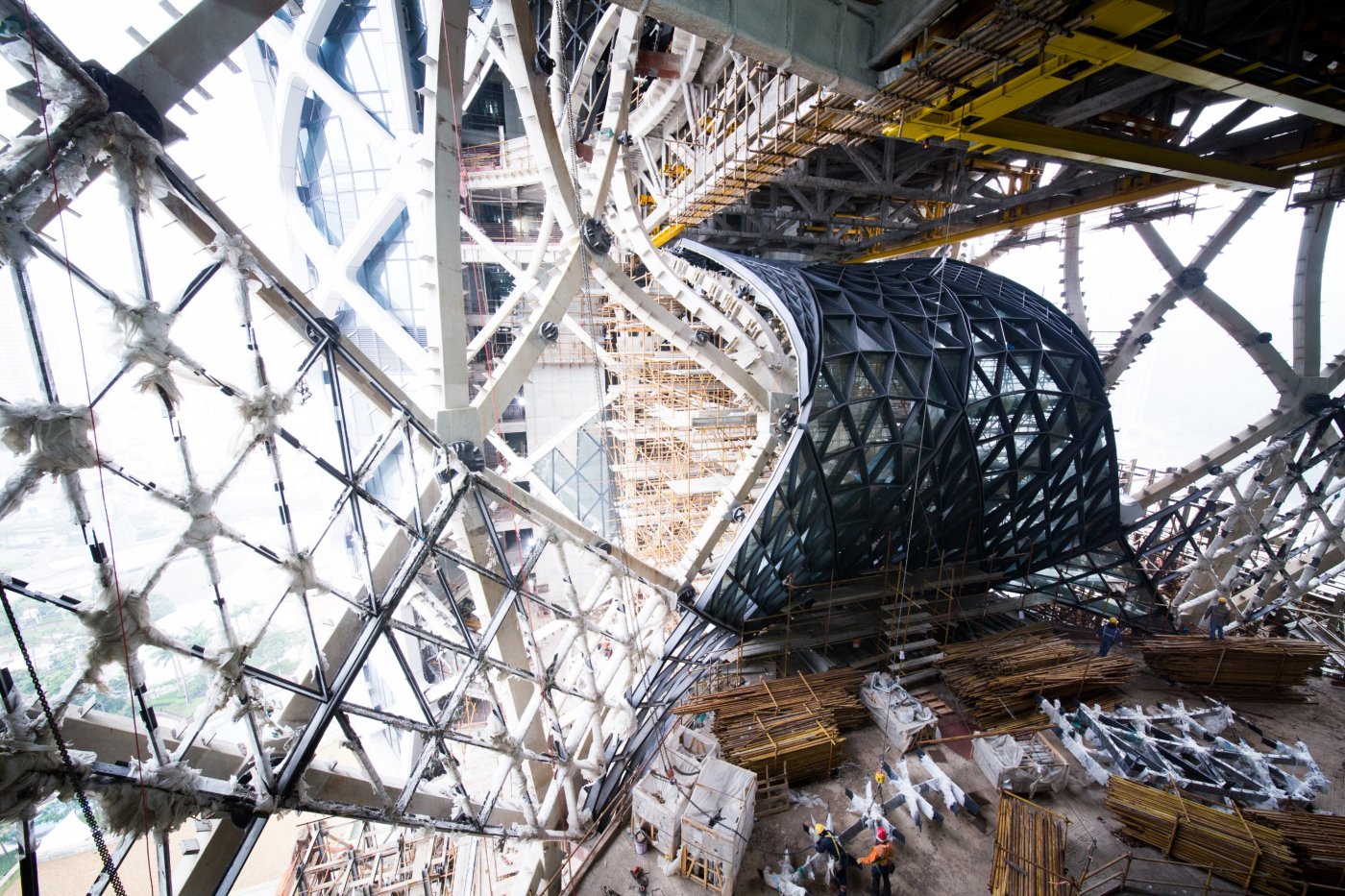 Looking up at the unfinished triangulated exoskeleton from inside the atrium, with scaffolding and site debris